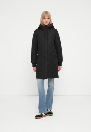 Schwarzer gesteppter Longcoat mit Kapuze, ausgestattet mit einem Frontreißverschluss und seitlichen Taschen. Trägt dazu hellblaue Schlagjeans und schwarze Sneakers.