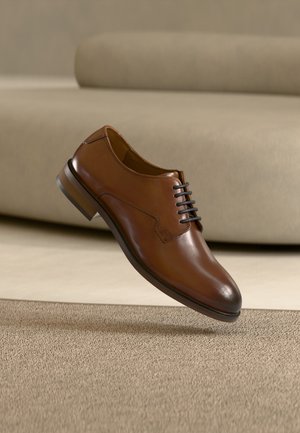 Καφετιά δερμάτινη επίσημη παπούτσια με μαύρα κορδόνια που αιωρείται πάνω από μπεζ χαλί σε ένα μινιμαλιστικό δωμάτιο με μια καμηλό καναπέ στο παρασκήνιο.