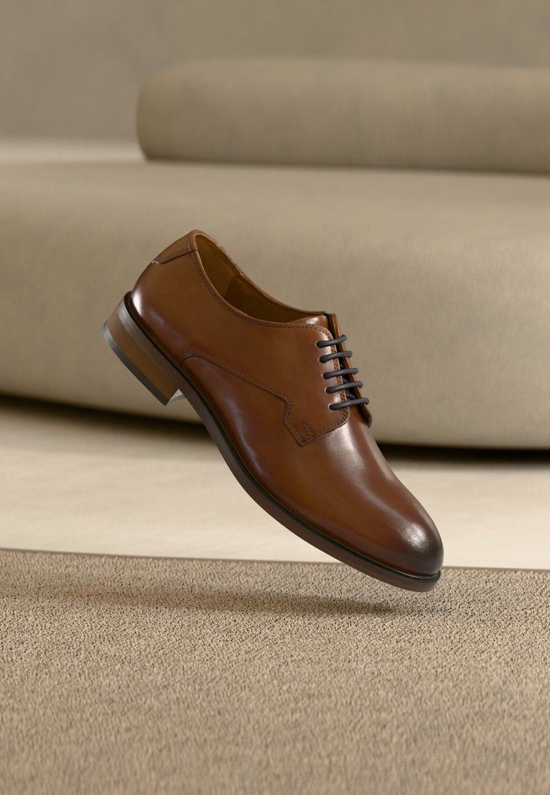 Καφετιά δερμάτινη επίσημη παπούτσια με μαύρα κορδόνια που αιωρείται πάνω από μπεζ χαλί σε ένα μινιμαλιστικό δωμάτιο με μια καμηλό καναπέ στο παρασκήνιο.