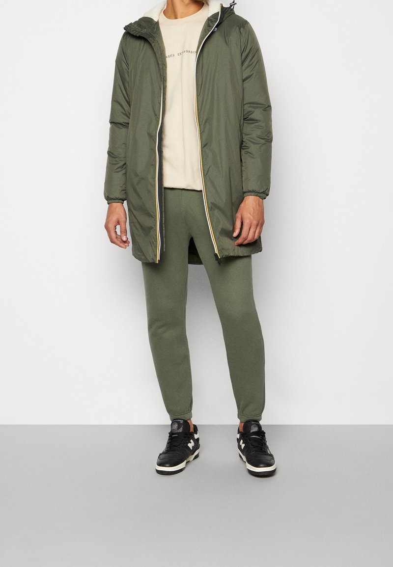 Giacca imbottita verde oliva con cappuccio e cerniera, felpa beige, pantaloni della tuta verdi abbinati e sneaker nere con dettagli bianchi.