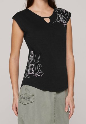 Czarna bawełniana koszulka z dekoltem w kształcie klucza, z rękawami typu cap, z nadrukowanym tekstem w różowym i białym, w połączeniu z zieloną spódnicą cargo.