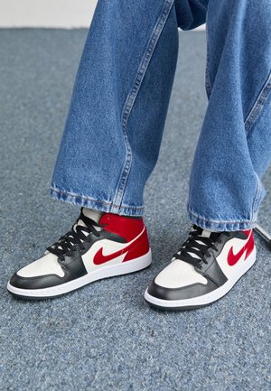 Stopy w czarno-biało-czerwonych butach Nike z czarnymi sznurówkami, w połączeniu z niebieskimi dżinsami, stojące na szarym wykładzinie.