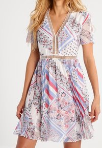 Vestido de manga corta con escote en V, que presenta una base blanca y coloridos patrones florales y geométricos en rojo, azul y púrpura, con una cintura ajustada.