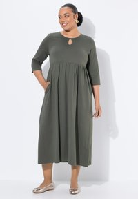 Olivgrön klänning i knälängd med trekvartsärmar, nyckelhålsknäppning, empiremidja och sidfickor; kombinerad med metalliska ballerina skor.