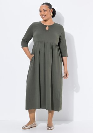 Olijfgroene jurk tot de knie met driekwart mouwen, keyhole-halslijn, empire taille en zijzakken; gecombineerd met metallic flats.