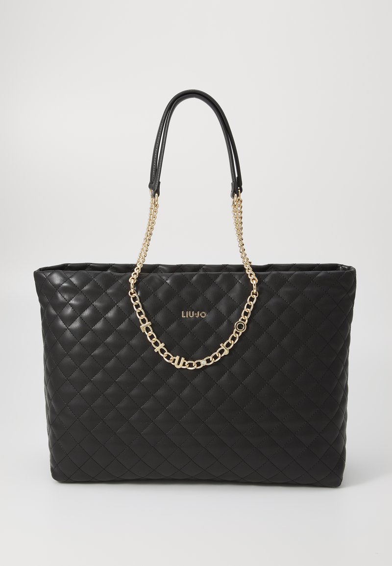 Sac tote noir matelassé en matériau synthétique, doté de deux poignées noires et d'une accentuation en chaîne dorée. Logo affiché de manière proéminente sur le devant.