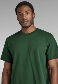 T-shirt verde a collo rotondo realizzata in cotone, con maniche corte e una superficie liscia. Design semplice senza motivi o decorazioni.