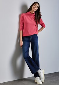 Rosa Rollkragenpullover aus weichem Material, kombiniert mit dunkelblauen, weit geschnittenen Jeans und weißen Turnschuhen mit cremefarbenen Sohlen.