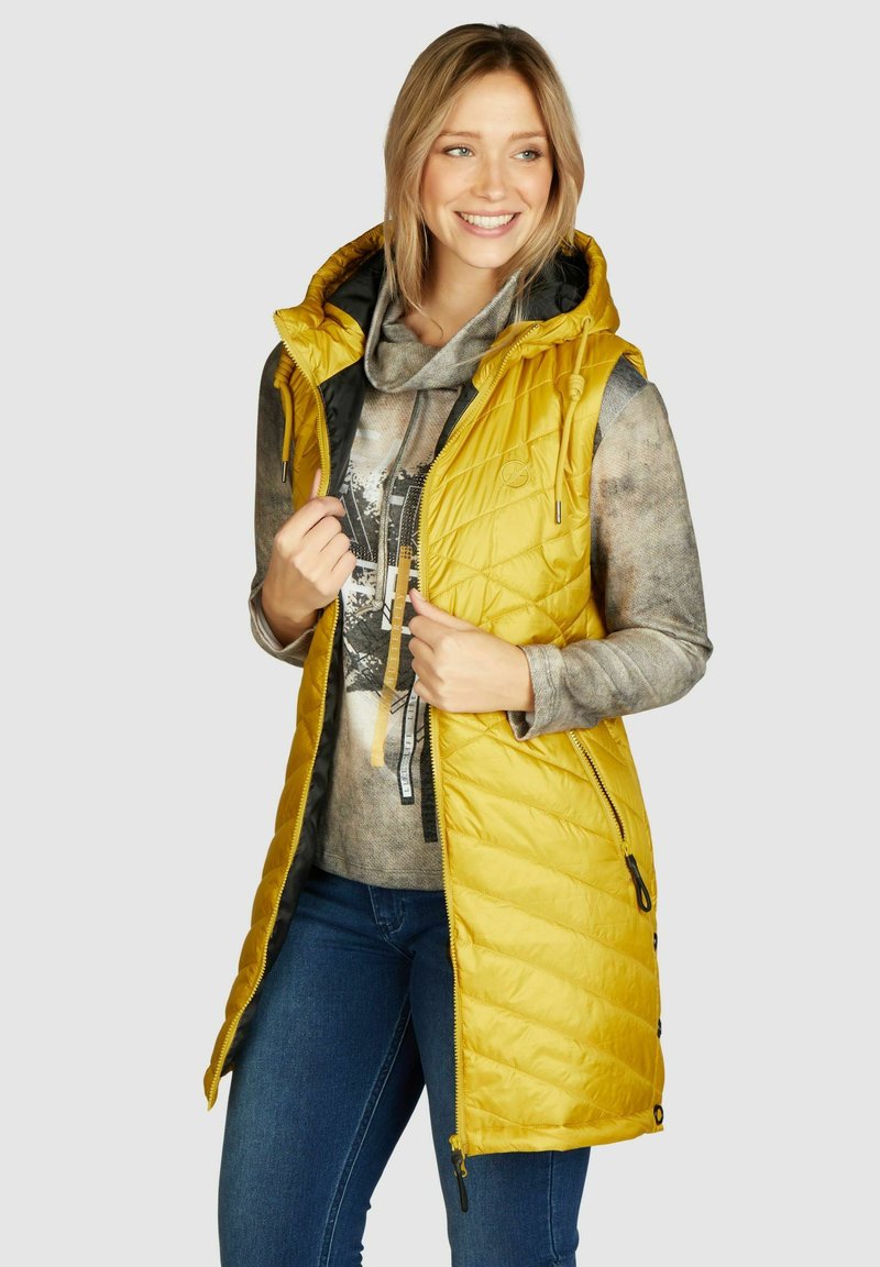 Geel gewatteerd vest met een capuchon, voorzien van ritszakken aan de voorkant en een zwarte voering. Gekozen boven een grijs patroon hoodie en een blauwe spijkerbroek.