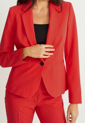 Czerwony, opięty żakiet i pasujące spodnie noszone na czarnej bluzce, z jedną ręką przytrzymującą żakiet na wysokości klatki piersiowej.