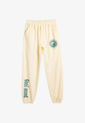 Pantalones de chándal de algodón color crema con cintura elástica, bolsillos laterales y gráficos verdes impresos en la pierna izquierda y en la parte trasera.