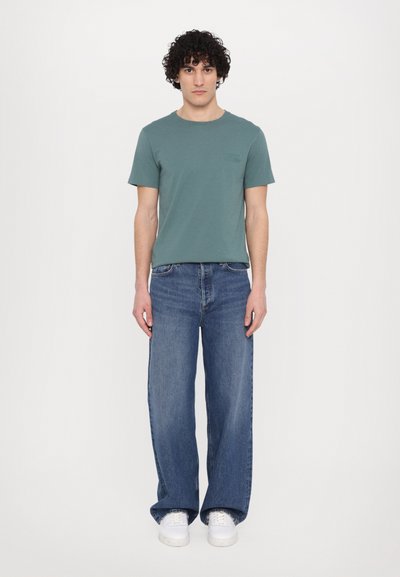 T-shirt en coton vert avec un col rond, associé à un jean bleu loose et des baskets blanches. Design simple, sans motifs visibles.