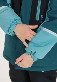 Türkis- und schwarze Jacke mit elastischen Bündchen, Reißverschluss-Taschen und reflektierenden Akzenten, aus wasserabweisendem Material gefertigt.