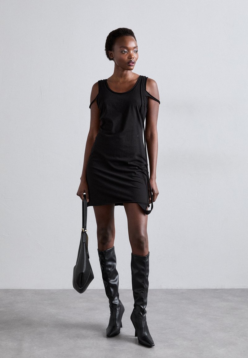 Schwarzes ärmelloses Kleid mit Ausschnitten an den Schultern, figurbetontem Design und seitlichem Schlitz. Tragen mit kniehohen schwarzen Lederstiefeln und einer passenden Handtasche.