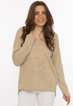 Lächelnde Frau mit langen braunen Haaren trägt einen beigen Pullover mit dem Text „Positive Mind Club“ und weiße Hosen vor einfarbig weißem Hintergrund.