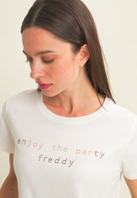 Camiseta de algodón blanca con mangas cortas, que presenta un diseño de texto colorido con puntos que dice "disfruta de la fiesta freddy" en la parte frontal.