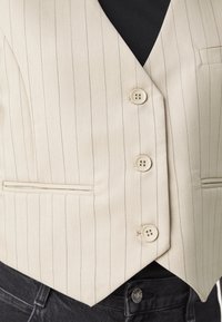 Gilet beige à fines rayures avec trois boutons porté sur un top noir et associé à un jean en denim noir.