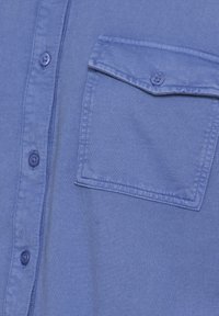 Camisa de botones azul claro con un bolsillo en el pecho, tejido texturizado y botones a juego. Detalle de costura visible en el bolsillo.
