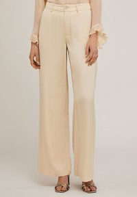 Breda byxor i mjuk beige satin, med en slät textur, en knappstängning och bälteshällor, kombinerade med guldiga högklackade sandaler med remmar.