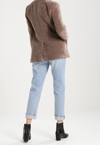 Osoba w jasnoniebieskich, podwiniętych dżinsach, czarnych botkach za kostkę i brązowym, teksturowanym płaszczu stoi na tle jednolitego tła.