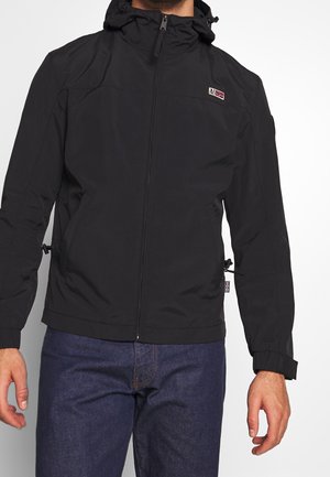 Mann trägt eine schwarze Kapuzenjacke mit Reißverschluss und Seitentaschen sowie einem kleinen Flaggenlogo auf der Brust, kombiniert mit dunkelblauen Jeans.