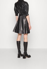 Robe en cuir noir avec une jupe plissée, des manches ajustées et une ceinture à nouer ; associée à des bottes noires en cuir jusqu'aux genoux avec des talons épais.