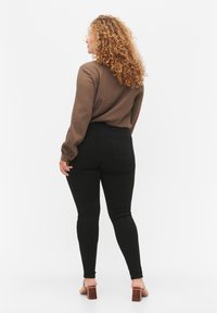 Top largo de manga larga texturizado en marrón combinado con jeans skinny negros de cintura alta. La modelo lleva sandalias de tacón bloque, vista desde atrás.