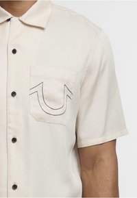 Camisa de manga corta beige hecha de algodón; presenta un bolsillo con un detalle de costura negra y botones negros en la parte delantera.