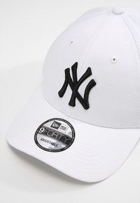 Valkoinen baseball-lippis, jossa musta brodeerattu "NY"-logo. Varustettu kaarevalla viseerillä ja säädettävällä hihnalla. Tunnusmerkki osoittaa "9FORTY"-tyylin.