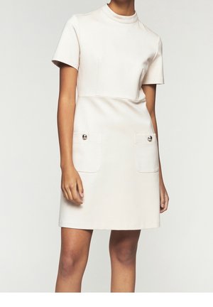 Femme portant une robe crème à manches courtes, longueur genou, avec deux poches avant ornées de boutons argentés.