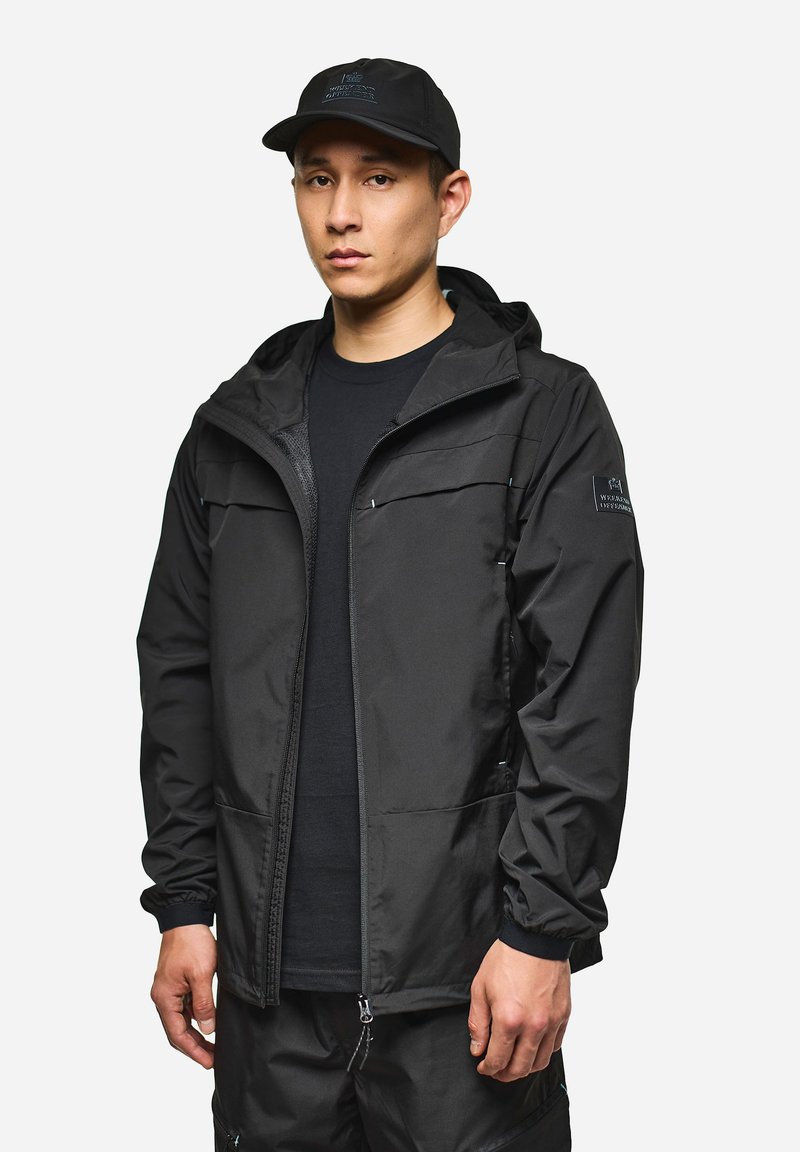 Giacca leggera nera con cappuccio, chiusura con zip frontale e polsini elasticizzati. Presenta due tasche sul petto e una toppa con logo sulla manica.