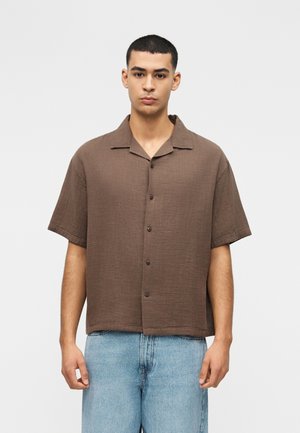 Νέος άνδρας με κοντά μαλλιά, που φοράει ένα φαρδύ καφέ πουκάμισο με κοντά μανίκια και ανοιχτό μπλε τζιν, στέκεται μπροστά σε ένα απλό φόντο.