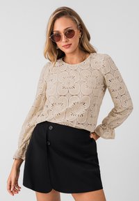 Beige kanten top met bloemenpatronen en lange mouwen, gecombineerd met een zwarte wikkelskirt met twee knopen voor sluiting.