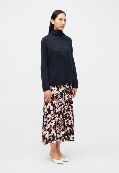 Μπλε σκούρο ριγωτό πουλόβερ με ψηλό γιακά, συνδυασμένο με φούστα μέχρι το πάτωμα με λουλουδάτο μοτίβο σε αποχρώσεις μωβ και λευκού. Άσπρα μυτερά φλατ παπούτσια.