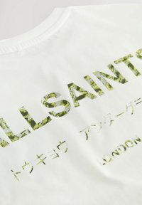 Valkoinen puuvillapaita, jossa on tekstuurilla varustettu vihreä graafinen teksti "ALLSAINTS" ja lisämerkkejä eri fonteilla painettuna selässä.
