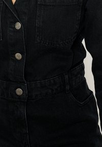 Črn jeans enodelni kombinezon s kovinskimi gumbi, dvema žepoma na prsih, pasom in teksturirano tkanino s rahlo zbledelim videzom.
