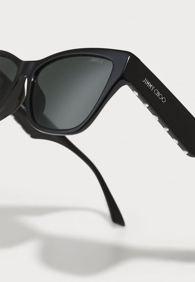 Ochelari de soare cu formă de ochi de pisică, fabricați din plastic lucios de culoare neagră, cu lentile gri. Prezintă accente cu cuie argintii și branding gravat pe braț.