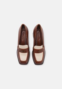 Mocasines de cuero marrón con puntera cuadrada, detalles en cuero crema, una correa decorativa y un tacón bajo. Suela texturizada con detalles de costura.