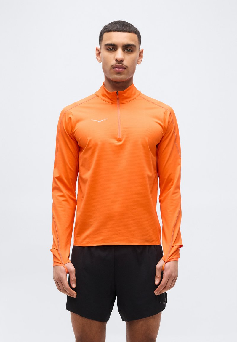 Άνδρας που στέκεται μπροστά φοράει πορτοκαλί αθλητική μπλούζα μακρυμάνικη με τρύπες για τον αντίχειρα και μαύρο σορτς, πάνω σε απλό φόντο.