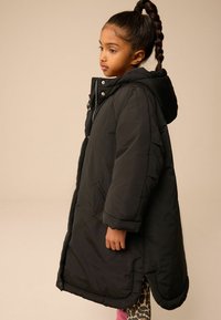 Cappotto piumino nero con cappuccio, dotato di bottoni a pressione, tasche laterali e vestibilità rilassata. Tessuto liscio con orlo fino al ginocchio.
