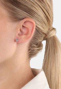 Kvinna med blont hår i hästsvans som bär en liten glänsande silvrig kula som örhänge på sitt vänstra öra mot en enkel bakgrund.