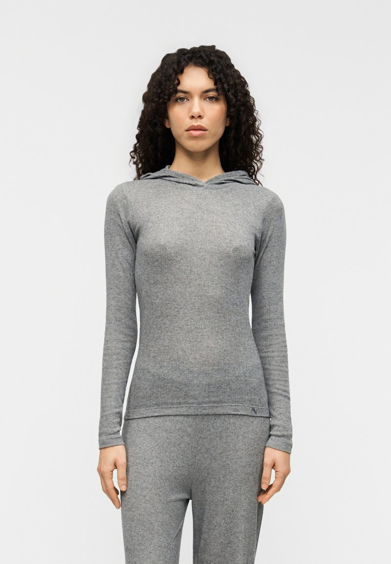 Γκρι μπλούζα με μακριά μανίκια και κουκούλα, κατασκευασμένη από απαλό, υφασμάτινο ύφασμα με υφή. Διαθέτει στρογγυλή λαιμόκοψη, εφαρμοστή γραμμή και διακριτικό λογότυπο της μάρκας κοντά στο τελείωμα.