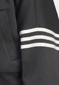 Manica di felpa nera con tre righe orizzontali bianche parallele e dettaglio di tasca a marsupio sul lato.