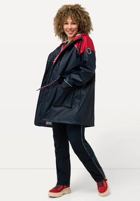 Parka imperméable bleu marine avec des accents rouges, capuche avec cordon de serrage et poches. Associée à un pantalon noir et des chaussures de randonnée rouges.