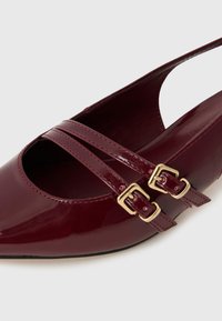 Burgundy kiillotettu nahka slingback-kengä, jossa on teräväkärkinen muotoilu, kahdella ristiin menevällä hihnalla ja kultaisilla soljilla säätöä varten.