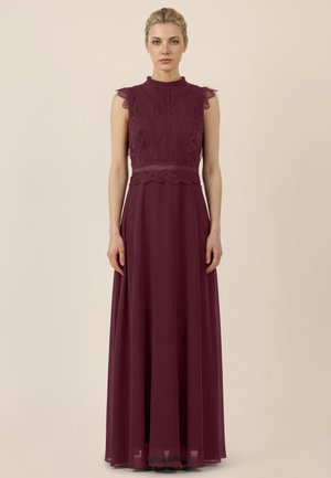 Frau steht aufrecht und trägt ein ärmelloses, burgunderfarbenes Spitzenoberteil und ein bodenlanges Chiffonrockkleid vor einem schlichten Hintergrund.