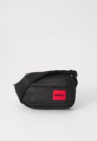 Geantă crossbody din țesătură neagră, cu un patch roșu cu logo, formă rectangulară, compartimente cu fermoar și curea ajustabilă. Suprafață texturată.