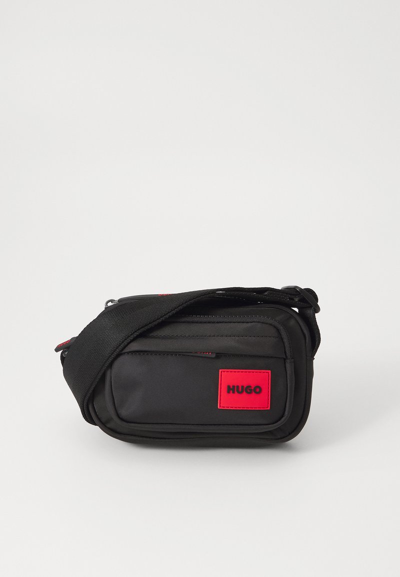Geantă crossbody din țesătură neagră, cu un patch roșu cu logo, formă rectangulară, compartimente cu fermoar și curea ajustabilă. Suprafață texturată.