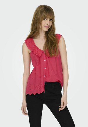 Roze mouwloze blouse met een gegolfde kraag, gaatjesdetail en knoopsluiting, gecombineerd met zwarte broek, tegen een lichte achtergrond.