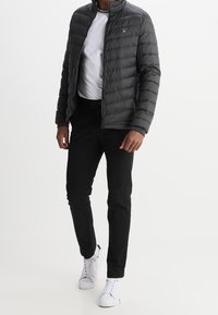 Miestnas pilkas pūstas švarkas su aukštu apykakle, užtrauktuku priekyje, dėvimas ant balto marškinio, derinamas su juodomis kelnėmis ir baltomis sportinėmis bateliais.